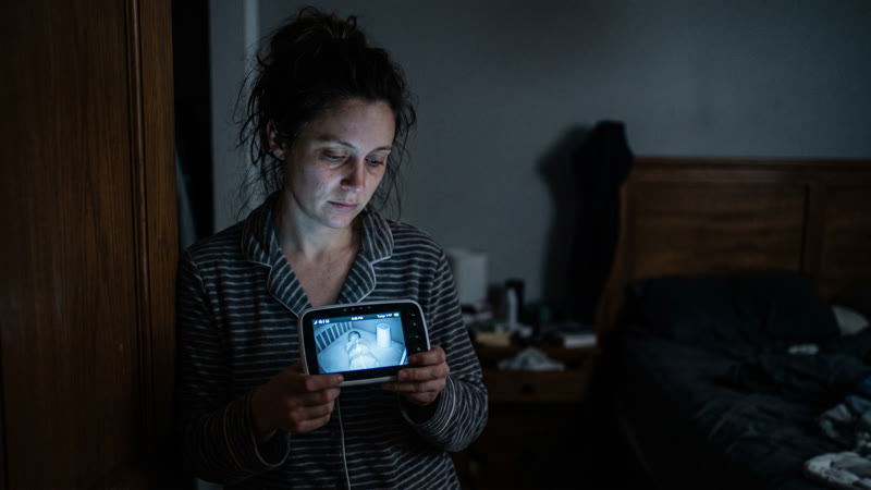 Tired parent in pajamas checking a baby monitor in a dark bedroom at night — baby not sleeping through